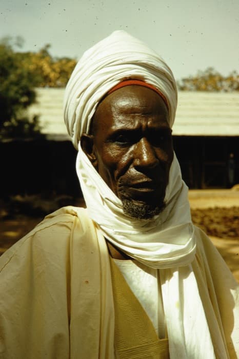 A man in a white turban, photographed outside a market shed. Northern Nigeria, 1961.